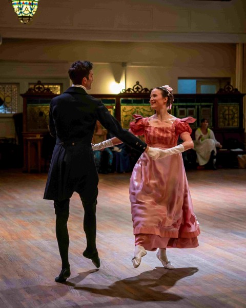 A man wearing a black suit and a woman wearing a pink dress and white gloves hold hands as they dance.