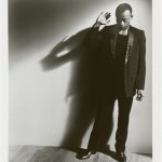 A man wearing a black tuxedo jacket and pants stands against a wall with head down and right arm raised.