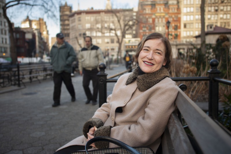 A smiling woman wearing a brown coat sitting on a bench.