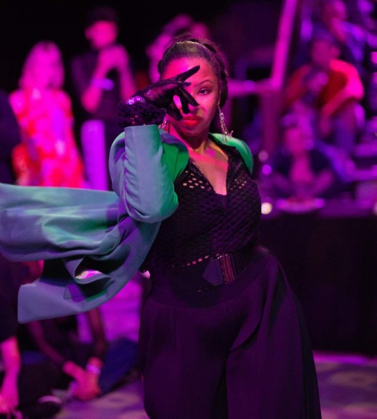 A woman poseswith her right arm bent and gloved hand in front of her face.