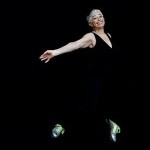 A woman wearing black stands in a tap dance pose with her arms outstretched