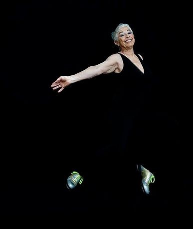 A woman wearing black stands in a tap dance pose with her arms outstretched