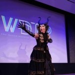 A performer wearing a black dress, black gloves and black headpiece stands extending their right arm out.