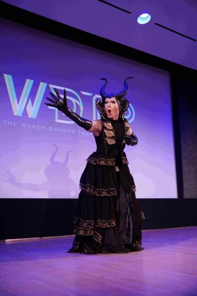 A performer wearing a black dress, black gloves and black headpiece stands extending their right arm out.