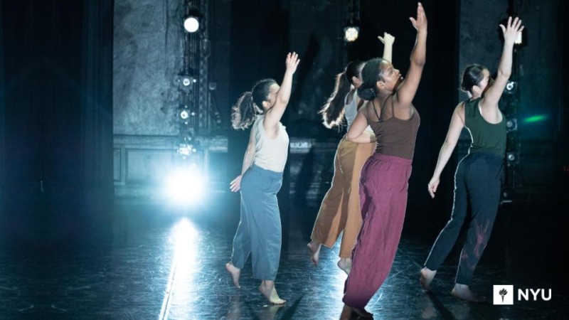 Four dancers in multicolored clothing reach their right arm up with NYU logo on the bottom right corner