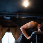 Male-Identifying dancer in front of a curtain, leaning on a black desk.