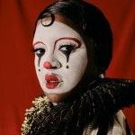 Joy-Marie Thompson, a Black person is seen dressed up as JoyBoy da Clown donning white face/ red nose makeup pensively looks on.