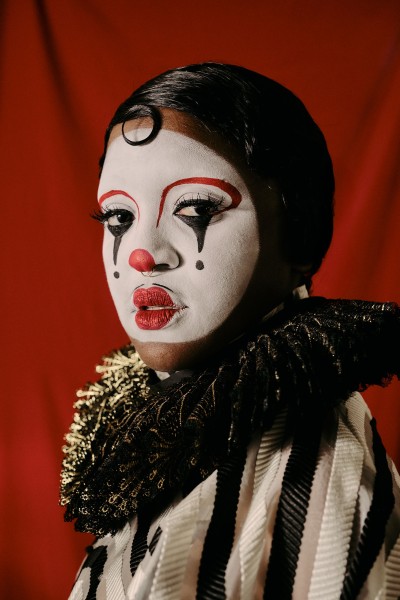Joy-Marie Thompson, a Black person is seen dressed up as JoyBoy da Clown donning white face/ red nose makeup pensively looks on.