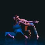 Two contemporary dancers sit on a dark stage floor under dramatic blue and pink lighting. Their left arms are reaching side.