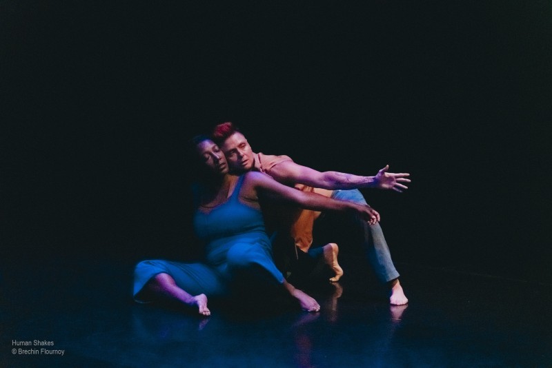 Two contemporary dancers sit on a dark stage floor under dramatic blue and pink lighting. Their left arms are reaching side.