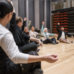 Participants sitting on the floor in a partial circle with one arm stretched upwards