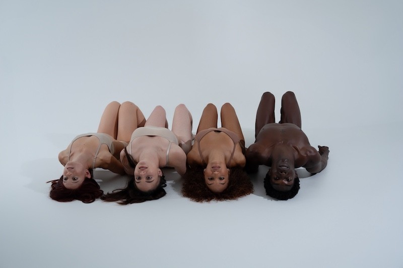 Four dancers of all shades are lying on the floor next to each other with their backs arched and eyes focused at the camera.