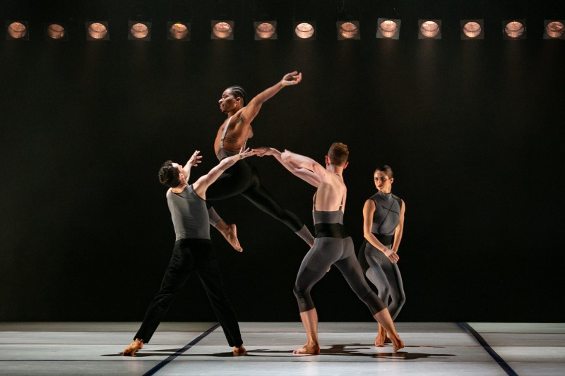 Contemporary dancers performing a dynamic lift on a dark stage, with one dancer airborne while others support and frame the move