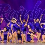 A group of young dancers in matching blue costumes pose together on stage during a dance competition, performing synchronized ch