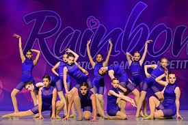 A group of young dancers in matching blue costumes pose together on stage during a dance competition, performing synchronized ch