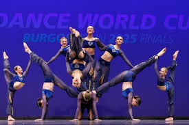 A group of competitive dancers perform an acrobatic formation on stage, with multiple dancers balancing upside down and lifting 