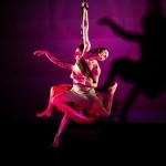 Two intertwinde women gracefully hang from a rope, lit in red, casting a stark shadow.