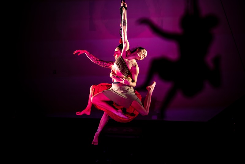Two intertwinde women gracefully hang from a rope, lit in red, casting a stark shadow.