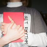 Person wearing layered shirts (a white long sleeve under a gray tee) holds a bright red notebook against their chest. Their hand