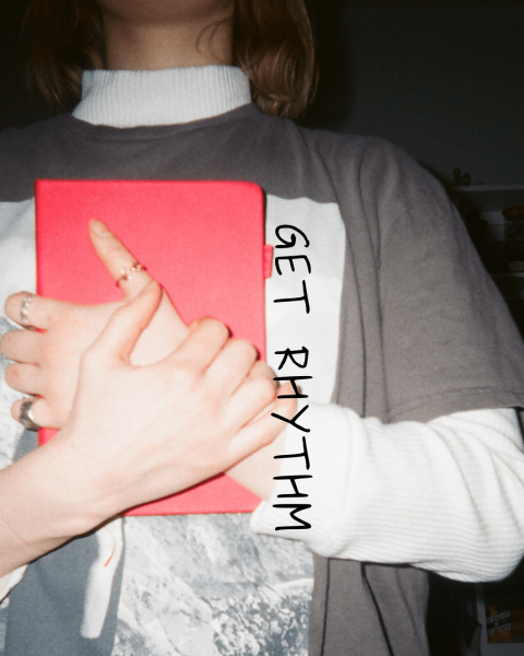 Person wearing layered shirts (a white long sleeve under a gray tee) holds a bright red notebook against their chest. Their hand