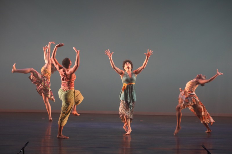 Four women dance and look up at the sky with their arms above their heads, palms open towards the ceiling.