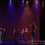 Seven dancers in solid-colored bodysuits stand facing the right with one hand outstretched to the audience.