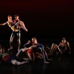 a group of seven dancers in strange poses looking at an imaginary TV