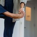Black person wearing navy and green standing on one leg, left arm holding right big toe across the center of their body.