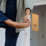 Black person wearing navy and green standing on one leg, left arm holding right big toe across the center of their body.