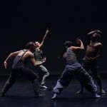 four dancers in dark clothing perform grounded movement on a low lit stage