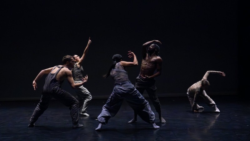 four dancers in dark clothing perform grounded movement on a low lit stage