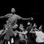 dancers with arms raised gather around a dancer sitting on another's shoulder 