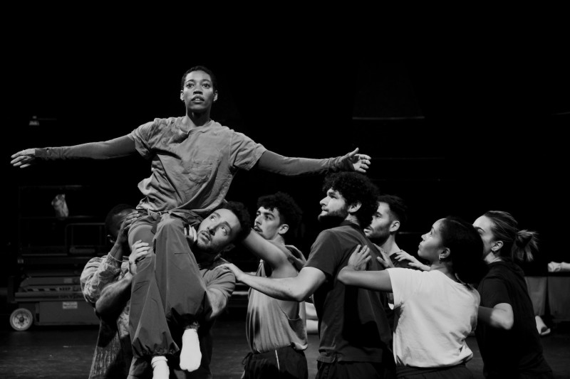 dancers with arms raised gather around a dancer sitting on another's shoulder 