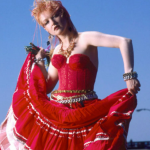 Photo of Cyndi Lauper in a bright red dress. Her head is tilted to the right and she is holding up the edges of her dress.