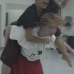 In a blurred image, a dancer wearing dark clothes leaps onto a second dancer, limbs wrapping around the other dancer 