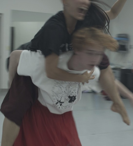 In a blurred image, a dancer wearing dark clothes leaps onto a second dancer, limbs wrapping around the other dancer 