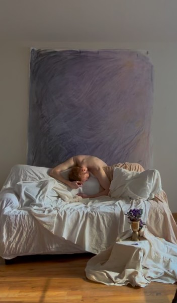 Male dancer slumped over an all white couch with a lavender backdrop and violets on the floor.