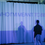Dancer stands with back to camera facing a white curtain with the words "WHO I'M MEANT TO BE" projected in purple