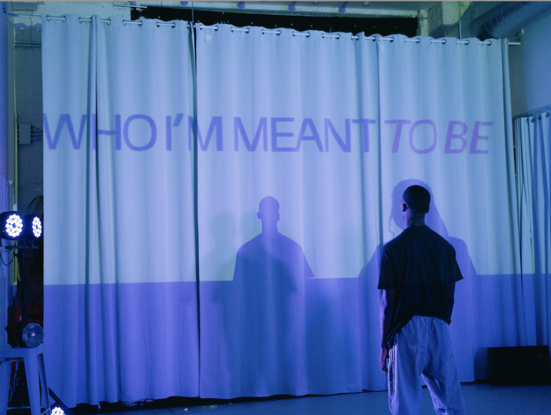Dancer stands with back to camera facing a white curtain with the words "WHO I'M MEANT TO BE" projected in purple