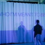 Dancer stands with back to camera facing a white curtain with the words "WHO I'M MEANT TO BE" projected in purple