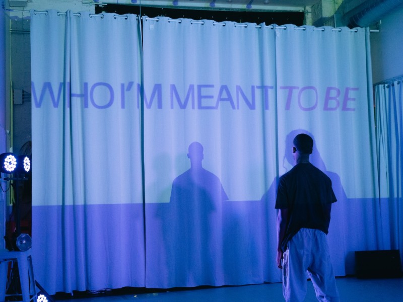  Dancer stands with back to camera facing a white curtain with the words "WHO I'M MEANT TO BE" projected in purple