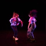 Two dancers face one another, lit in a pink light. They smile, kicking a leg out toward one another, performing a house duet.