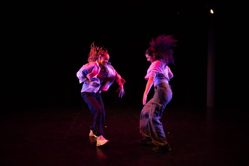 Two dancers face one another, lit in a pink light. They smile, kicking a leg out toward one another, performing a house duet.