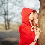 Girl with red hood, hiding behind a tree, looks out of the frame with a look of curiosity. Blurry trees in the background.