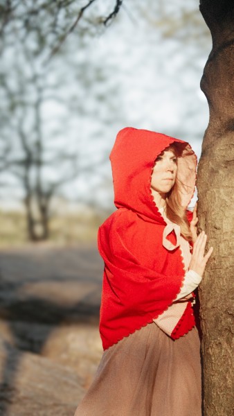 Girl with red hood, hiding behind a tree, looks out of the frame with a look of curiosity. Blurry trees in the background.