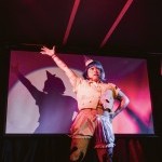 A performance photo of radio illuminated by spotlight. They have one arm raised in the air, and the other arm on their hip
