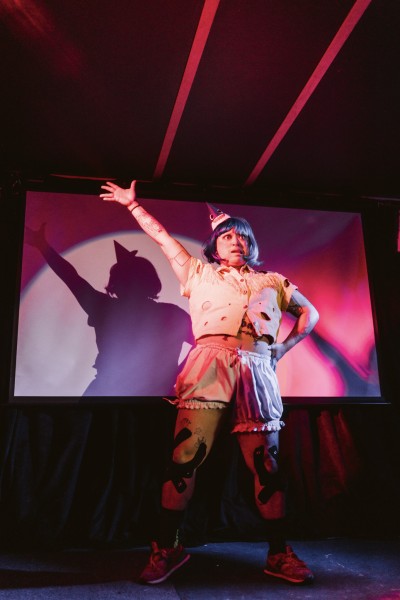 A performance photo of radio illuminated by spotlight. They have one arm raised in the air, and the other arm on their hip