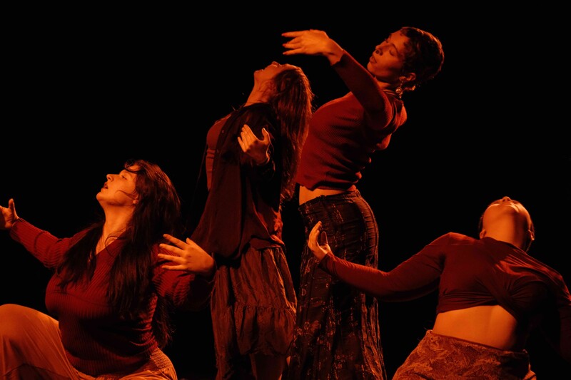 3 dancers dressed in red and brown lifting their chests to the sky with palms out and open, luxuriating in the movement