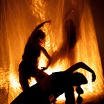 Three dancers appear in silhouette against a glowing amber-orange backdrop. Sheer, translucent fabric hangs from above, illumina