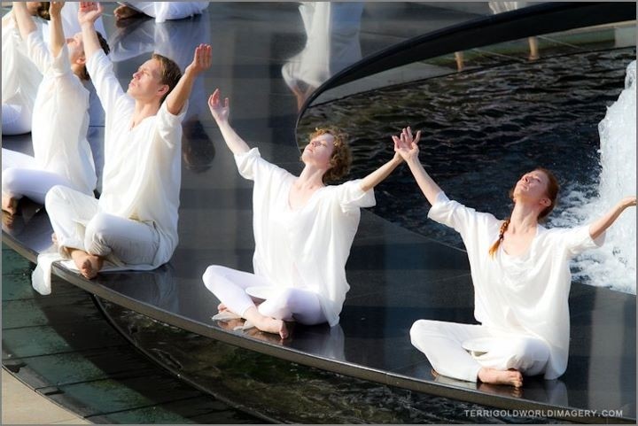 Dancers in white tops & leggings, sitting crossed legged on black fountain with arms raised to the sky